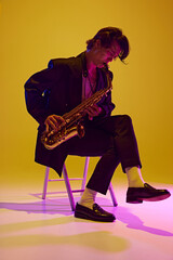 Fototapeta premium Young man, talented, charismatic solo player in black suit playing sax in neon light against vibrant yellow studio background. Concept of music and art, hobby, concerts and festivals, modern culture.