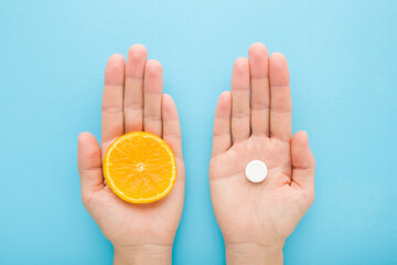Young adult woman hands holding white pill of c vitamin and cut slice of fresh orange fruit on pastel blue table background. Receiving nutrition supplement. Closeup. Point of view shot. Top down view. - 783033218