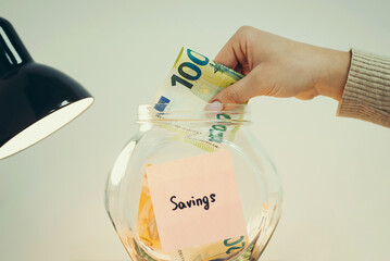 Word "Savings" written on a sticker, hanging on the glass money box, hand of a person putting euro banknote in it, close up, toned. Concept of savings, funding, investing and finances