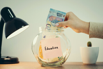 Glass jar with money standing on the table, female hand putting 20 euro banknote inside, lamp and kaktus nearby. Toned close up photo with concepts of saving funds for education