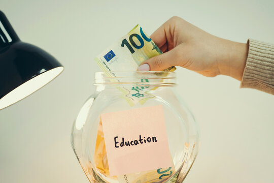 Pink sticker with word Education, hanging on the glass money box, female hand puts 100 euro banknote in it, close up, toned. Concept of saving money for knowledge and self development