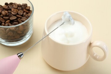 Mini mixer (milk frother), cup of whipped milk and coffee beans on beige background, closeup