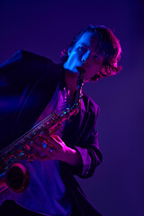 Young virtuoso in classical black suit playing saxophone in vibrant pink neon light against gradient studio background. Concept of music and art, hobby, concerts and festivals, modern culture. Ad
