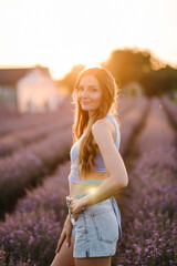 A young and slim girl walks and enjoying fun the time. Smiling female walking among lavender flowers with sunlight on summer day. Woman standing in lavender field and sniffing flowers at sunset.