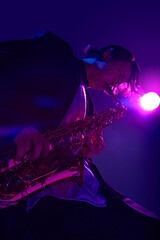 View from below of young talented man playing sax in vibrant pink neon light against dark studio...