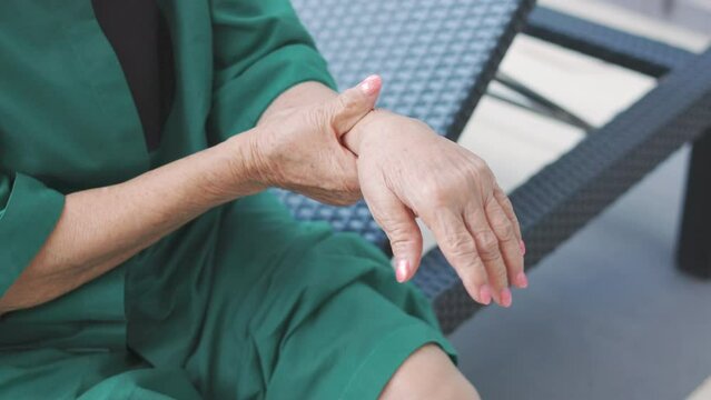 Close up 4K of hands holding painful wrist and arms of senior woman who is sitting on chair outdoor shows concept of health care problem of elderly people which needs social support for joint surgery.