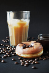 Caramel donut with coffee decor next to a glass of frappuccino with ice and a glass scoop of coffee on a dark background