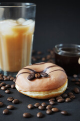 Caramel donut with coffee decor next to a glass of frappuccino with ice and a glass scoop of coffee on a dark background