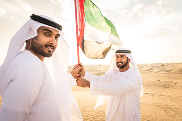 Two arab men wearing traditional emirati clothing in the desert of Dubai - Middle-eastern adult males portrait holding emirate flag to celebrate national day