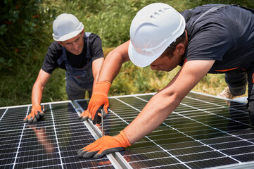 Male workers mounting photovoltaic solar panel system outdoors. Men engineers placing solar module on metal rails, wearing construction helmets and work gloves. Renewable and ecological energy.