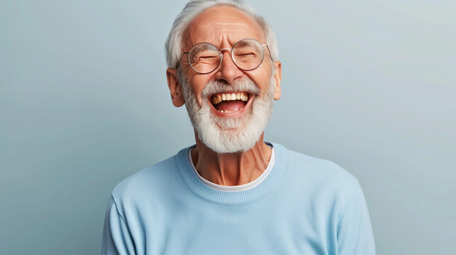 A man with a big smile on his face is wearing glasses and a blue shirt. He is laughing and he is happy. laughing old man, beard, glasses, happy, open laugh, enthusiastic, light blue sweater
