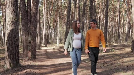 Happy delighted beautiful expectant parents wearing casual clothing walking in wood pregnant woman and man enjoying nature and fresh air looking asides with pleasure holding hands