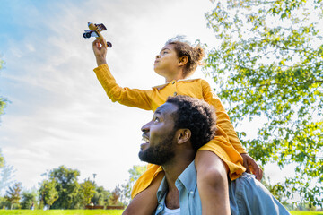 Beautiful happy african american family bonding at the park - Black father carrying on shoulders his lovely daughter and she is plaing with a toy airplane - obrazy, fototapety, plakaty