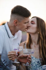 Male kisses female. Man hugs woman holds glasses of wine with lavender sprigs and drinks. Couple sits in lavender field on romantic evening at sunset. Straw basket and croissants on summer picnic.