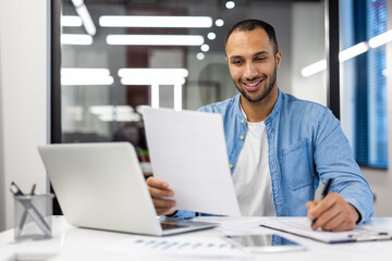 Fototapeta premium Young hispanic male businessman sitting in a modern office at a desk and working on a laptop and with documents