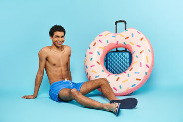 positive young african american man in swimming trunks sitting near suitcase and inflatable donut