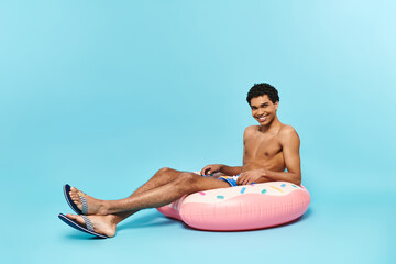 cheerful african american man in swimming trunks sitting on inflatable donut and smiling at camera