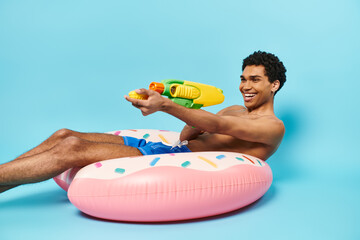 upbeat appealing african american man posing with water gun on inflatable donut on blue backdrop