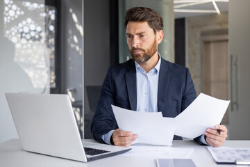 A serious young man businessman works in the office at a desk, looks at the screen of a laptop,...