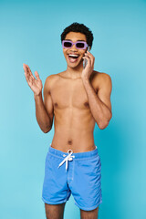 Fototapeta premium merry good looking african american man in swimming trunks with stylish sunglasses talking by phone