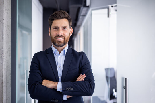 Portrait of a young successful businessman in a suit standing inside the office, crossing his arms on his chest, confidently and smilingly looking at the camera