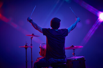 Rear view portrait of young artistic man playing drums in pink-purple stage lighting against...