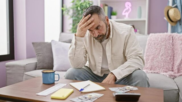 Mature bearded man looking stressed while examining bills at home, with money and coffee on table.