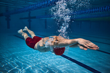 Dynamic image of athletic young guy, swimmer in cap in motion underwater, training in swimming pool. Bubbles, speed. Concept of professional sport, health, endurance, strength, active lifestyle - obrazy, fototapety, plakaty