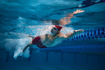 Dynamic image of young guy, swimming athlete in red cap and goggles in motion in swimming pool,...