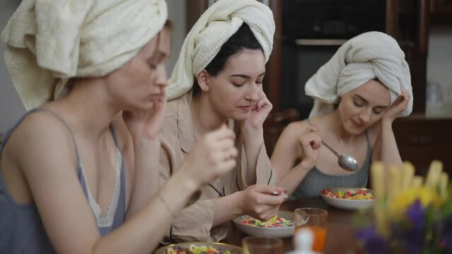 Three thoughtful young women with towels on their heads are having breakfast sitting at a table in the living room. Girls eat breakfast cereal with milk