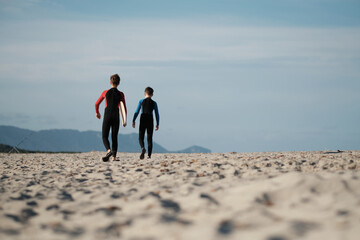 Junge Br&uuml;der im Neoprenanzug mit dem Skimboard am Strand auf Sardinien am Weg zum Wasser