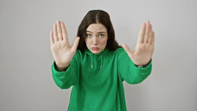Frustrated young woman, sweater clad and standing on a white isolated background, using hand gestures to signal 'stop', conveying annoyance and frustration.