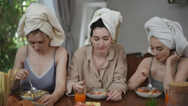 Three cute young women with towels on their heads are having breakfast while sitting at the table in the living room. Girls eat breakfast cereal with milk and look at the camera smiling