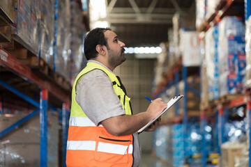 Male warehouse worker holding clipboard during working in storage warehouse. Warehouse worker inspecting quality of products on shelf pallet and checking stock inventory