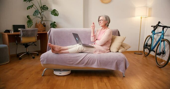 Timelapse video of mature old 60s woman student working at home using laptop technology device sitting on couch at home. While robot vacuum cleaner working around