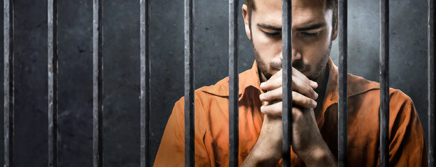 Young man praying behind bars in prison. Imprisonment and awaiting trial. Panorama with copy space.