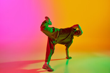 Young man, dressed streetwear dancing freestyle moves in motion in neon light against gradient pink-yellow background. Concept of art, hobby, sport, creativity, fashion and style, action. Ad