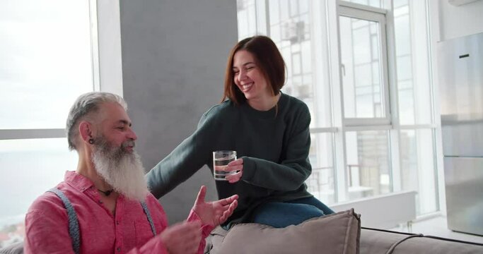 A happy brunette girl in a dark green jacket brings a transparent glass of water for her elderly father in gray hair and a pink shirt in a modern apartment