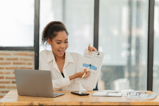 Young african american businesswoman displaying a financial chart on paper while talking during a virtual meeting or online presentation on her laptop from a modern office workspace