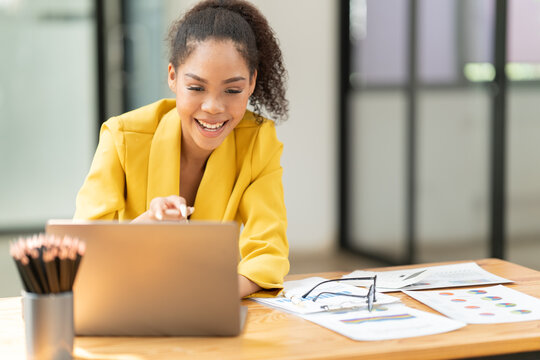 Confident young african american businesswoman smiling happily while working on a laptop, video calling, and participating in a virtual online meeting from her modern office or coworking space