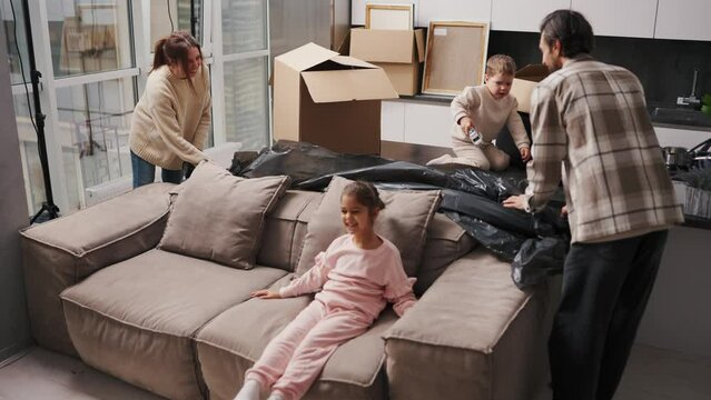 A happy brunette girl in a beige sweater together with her brunette husband in a plaid shirt lifts a black cover from the sofa and their little daughter jumps on it after moving to a new apartment