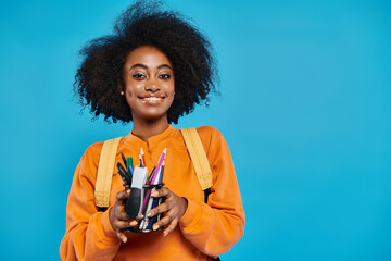 An African American college girl in casual attire holds a cup filled with an assortment of pens and...