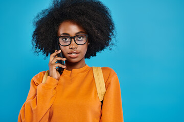 An African American college girl with glasses chats on a cell phone against a blue backdrop in a...