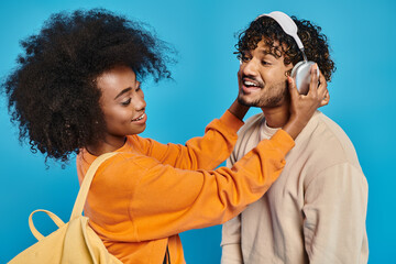 An interracial man and woman stand together wearing headphones against a blue backdrop, enjoying...