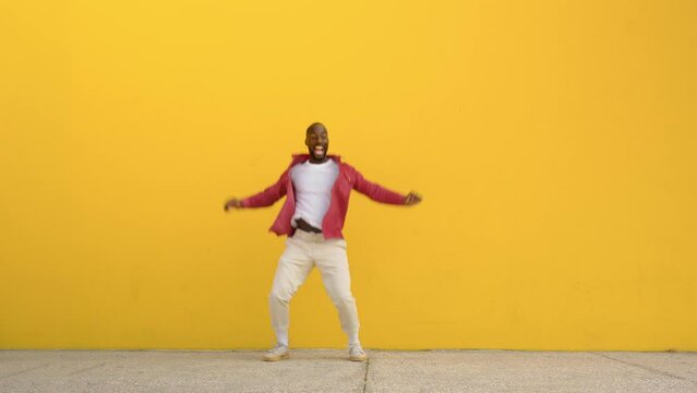 Happy excited cool stylish funky crazy young African Black man wearing red shirt feeling joy freedom having fun jumping in air celebrating win standing at yellow color urban wall background. Full body