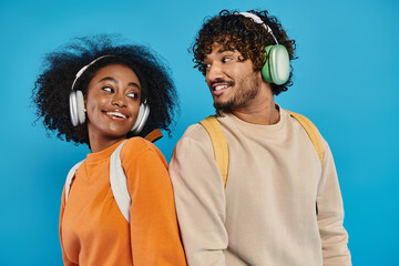 An interracial couple stands together, wearing headphones, sharing music on a blue backdrop in a studio setting. - obrazy, fototapety, plakaty
