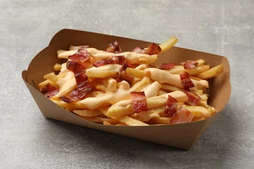 Poster Tasty potato fries, cheese sauce and bacon in paper container on grey table, closeup © New Africa