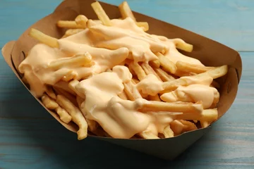 Poster Tasty potato fries and cheese sauce in paper container on light blue wooden table, closeup © New Africa