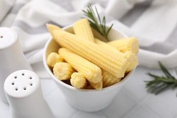 Tasty fresh yellow baby corns in bowl on white tiled table, closeup