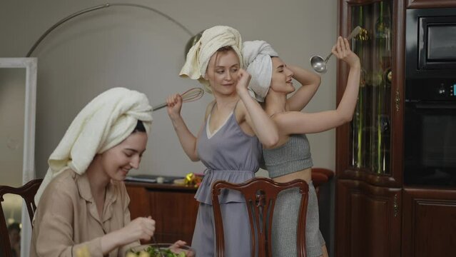 Two girls with towels on their heads dance with kitchen utensils in their hands in a modern kitchen, while a third girl sits at the table stirring a vegetable salad. Cheerful women are preparing